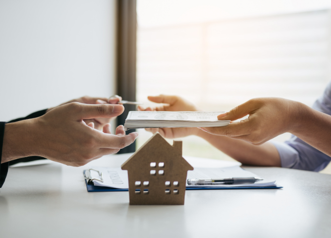 Two people exchanging documents over a table with a small house model in the foreground, representing a real estate transaction.