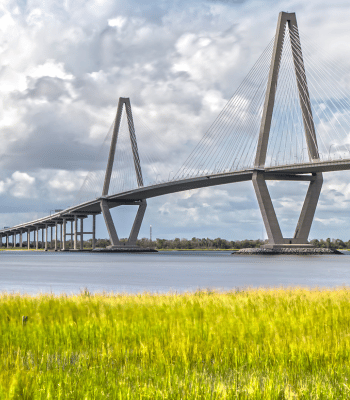 Scenic view of the Arthur Ravenel Jr. Bridge spanning over a lush green field and water in Mt. Pleasant, SC.